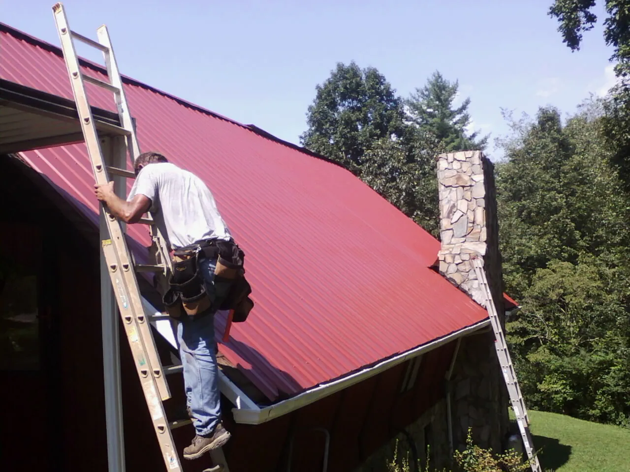 Roofer on a ladder beside a red metal roof and stone chimney — Soffit & Fascia Repair by Roofing Brothers in Forestville